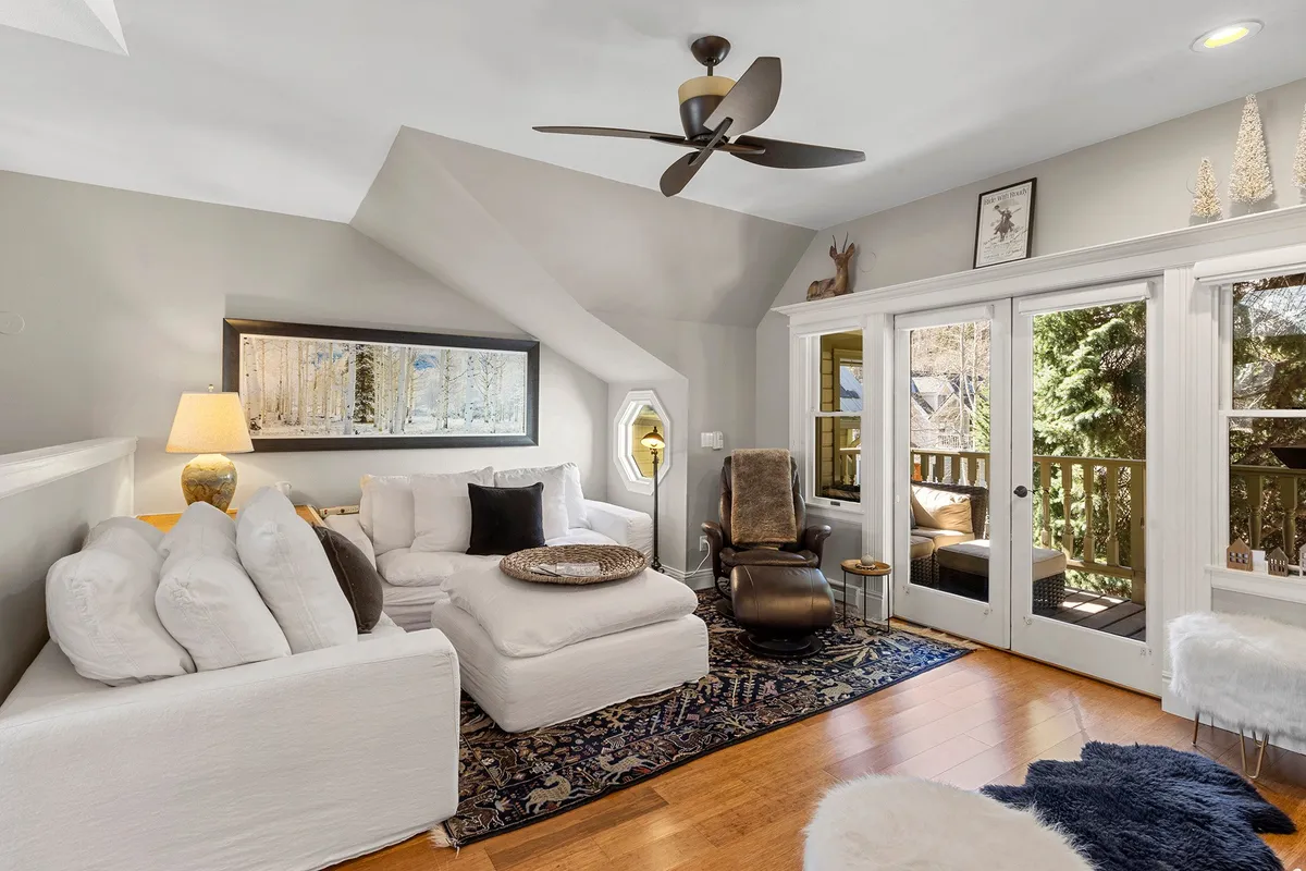 A bright, open living area featuring a plush white sectional sofa, decorative artwork on the walls, warm wooden floors, and large glass doors that lead out to a balcony. The space is filled with natural light, creating an inviting atmosphere ideal for relaxing, reading, or entertaining guests.