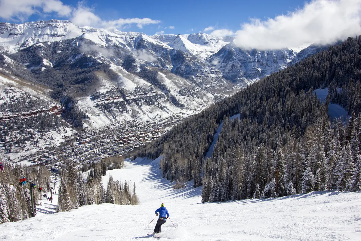 A skier navigates a pristine snow-covered slope, with dramatic mountains in the background and the Telluride town visible in the valley below. Evergreen trees frame the scene, emphasizing the close relationship between town and nature.