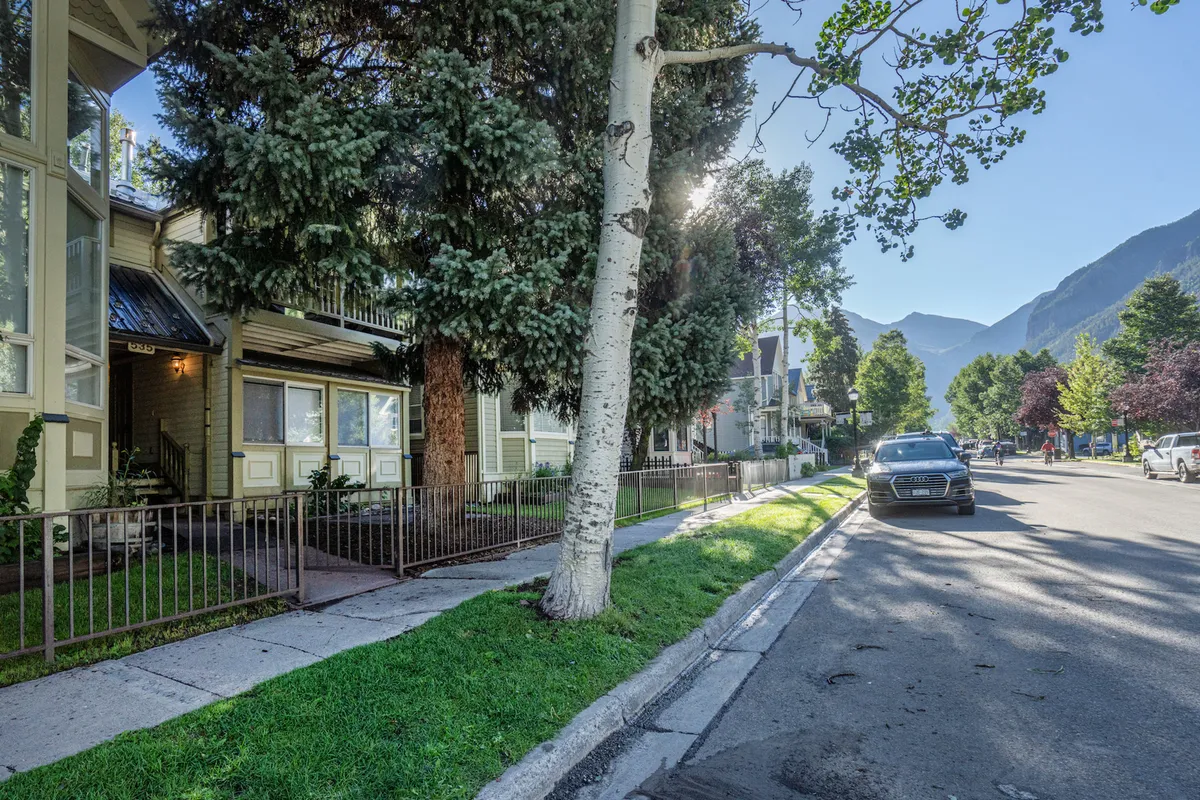 This exterior image shows a friendly front yard with green grass, tall trees, a sidewalk, and a parked car. The environment is residential and inviting, suggesting ease of access for walking or biking, and a peaceful setting for a mid-day break.