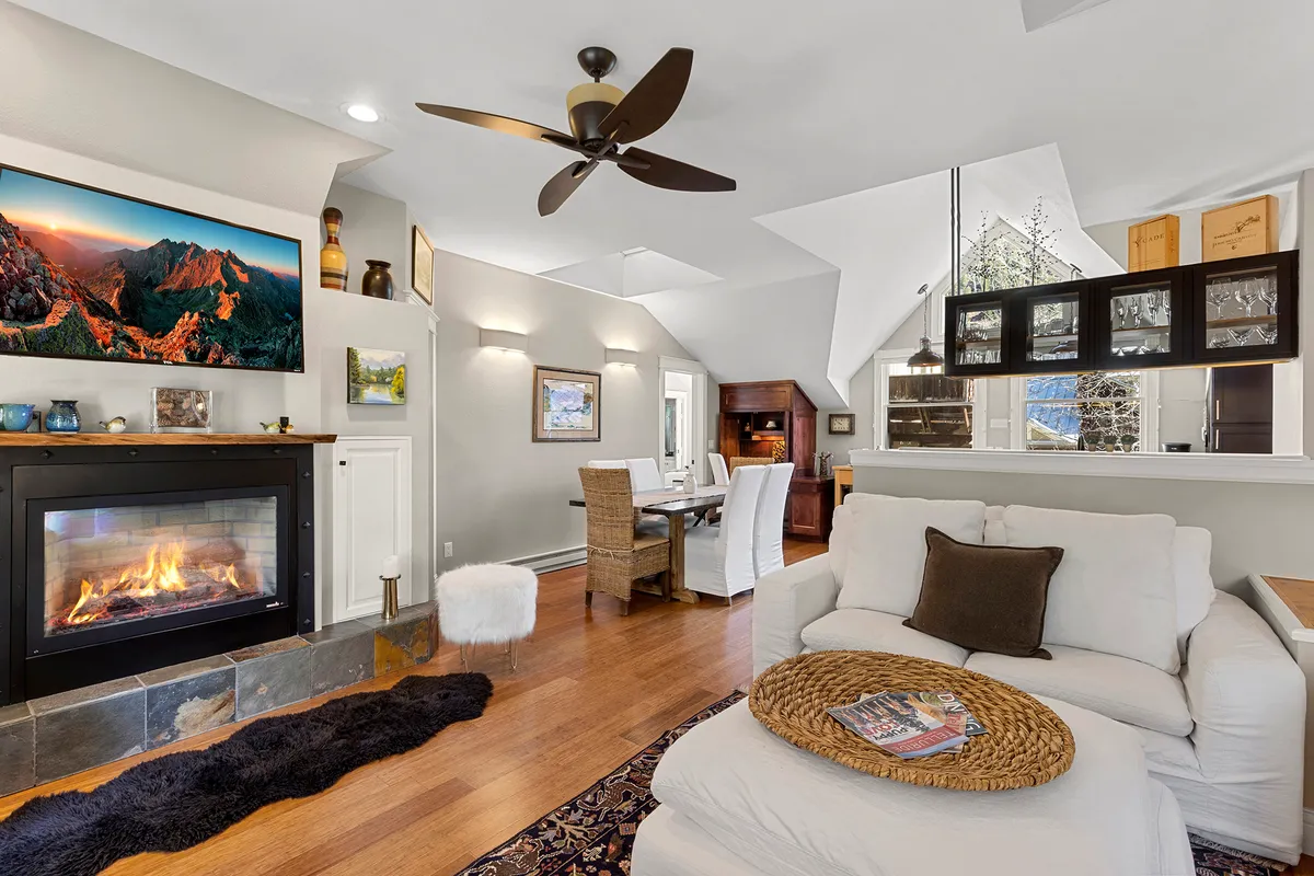 The image showcases a warm, inviting living area featuring a crackling fireplace, a plush white sofa, an elegant dining table set for gatherings, and tasteful artwork adorning the walls. The space is filled with natural light and exudes comfort, perfect for relaxing, entertaining, and enjoying time with family or friends.