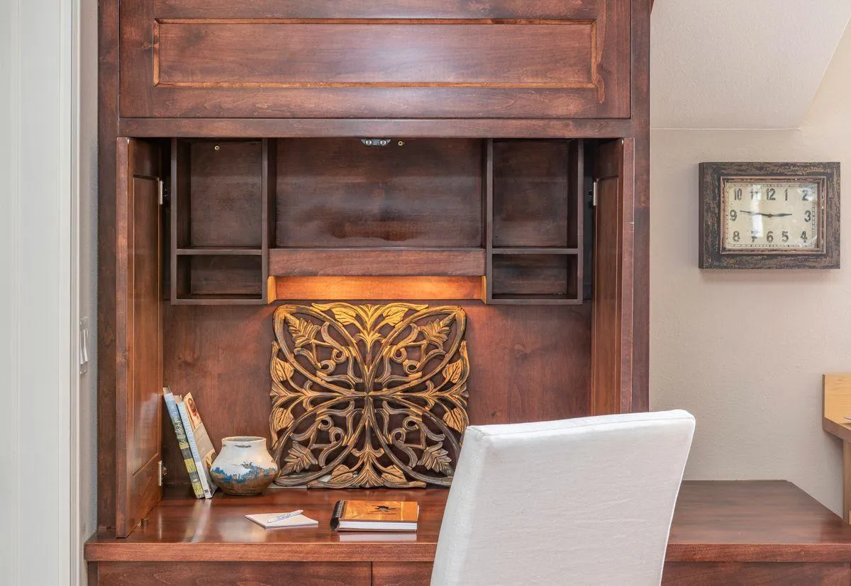 A cozy workspace featuring a wooden desk, a decorative pillow, shelves filled with books, and a wall clock. The desk is set apart from the main living areas, offering privacy and focus for work or study. Soft natural light and homey touches create an inviting, productive atmosphere.
