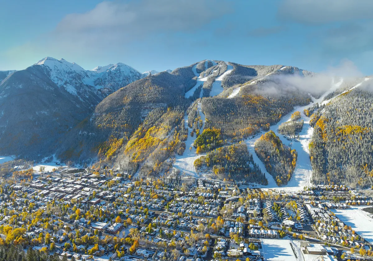 A panoramic view from the mountain peaks looking down onto snow-capped slopes and colorful autumn foliage with the charming town of Telluride nestled below. People can be seen skiing and snowboarding, while hiking trails snake through the vibrant landscape.