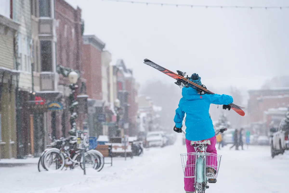 A playful winter scene featuring a person riding a bike with skis attached, navigating a snowy street lined with colorful buildings. The lively street scene hints at local shops and energetic community life, illustrating Telluride’s unique character and accessibility.