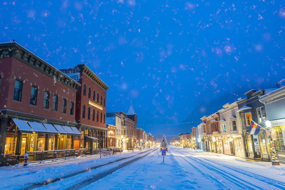 A charming, snow-covered street lined with historic buildings and decorated storefronts, featuring a Christmas tree and twinkling lights. Shoppers and visitors stroll along the festive sidewalks, creating a lively yet cozy winter atmosphere that captures the essence of Telluride’s downtown vibrant scene.