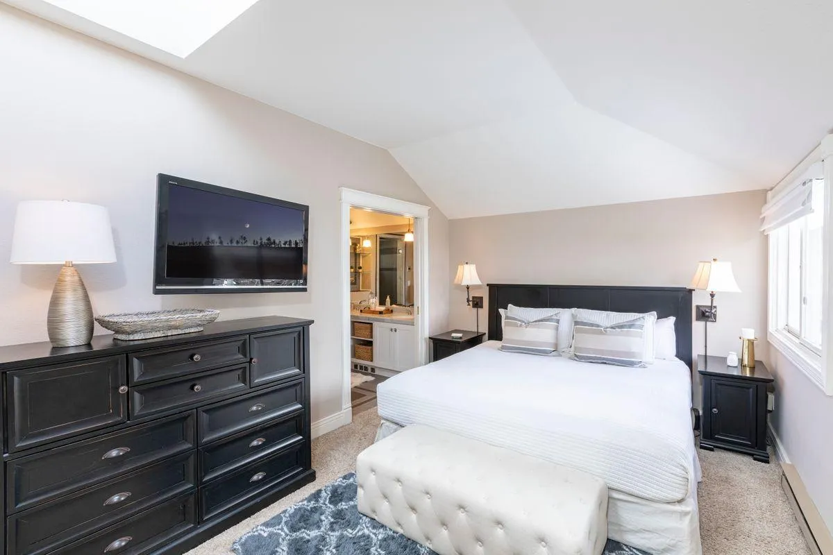 A serene master bedroom with a plush bed dressed in white bedding, a decorative ottoman at the foot, a modern lamp for warm lighting, and a flat-screen TV mounted on the wall. The window offers a glimpse of the outdoors, providing a tranquil escape.
