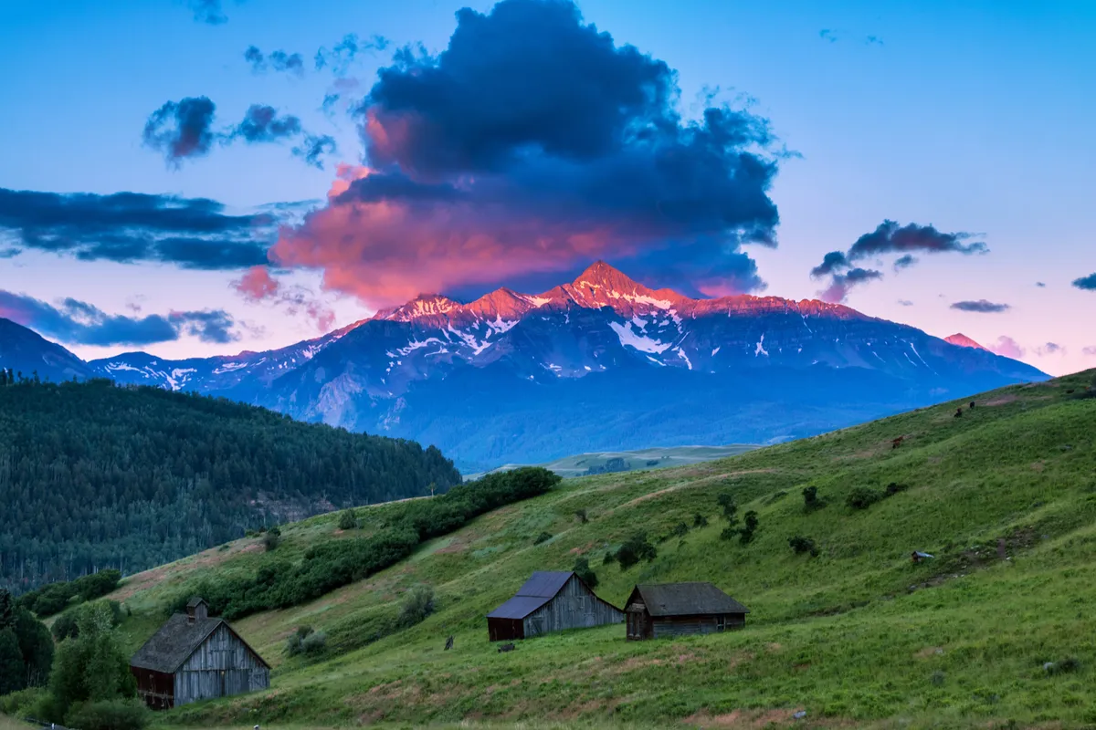 This wide-angle exterior shot features a sweeping mountain range under a dramatic sky, with verdant green hills and rustic cabins nestled at the base. The sense of open space and fresh air evokes the feeling of mountain breezes and relaxation on an elevated deck.