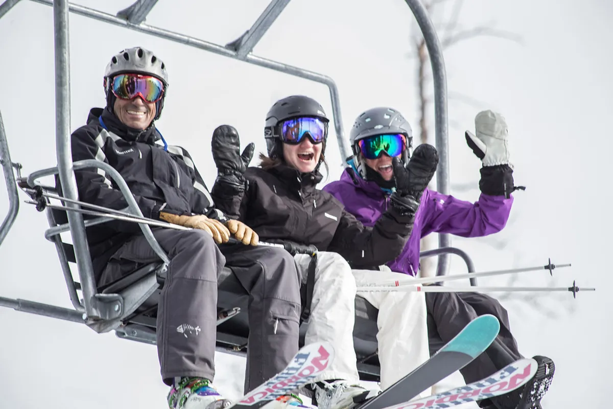 This image captures three skiers riding a chairlift above snow-covered slopes, with clear views of their ski gear and the expansive, snowy landscape in the background. The skiers are smiling and engaging with each other, conveying the excitement and camaraderie of winter sports in Telluride.