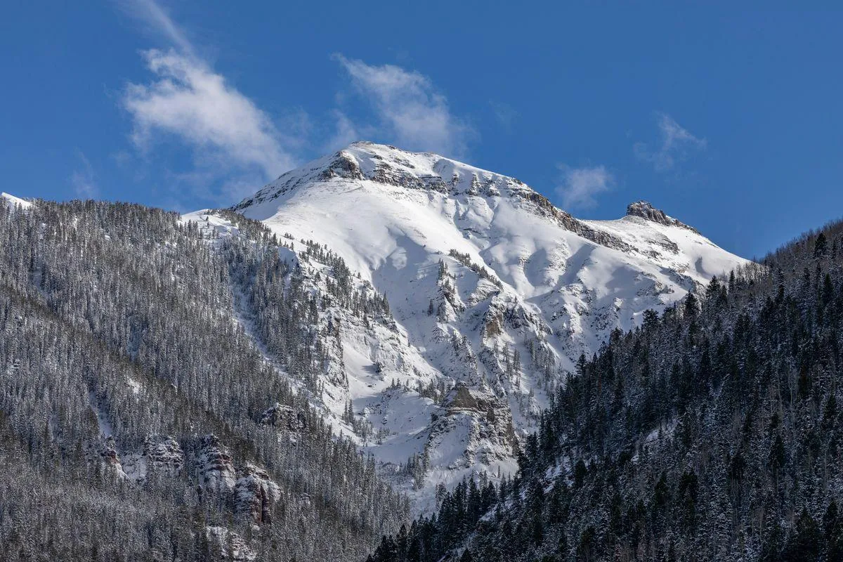 A tranquil view of a snow-capped mountain rising above a dense forest, set beneath a dramatic, cloudy sky. The scene invites thoughts of hiking, exploring hidden trails, and enjoying serene moments in nature.