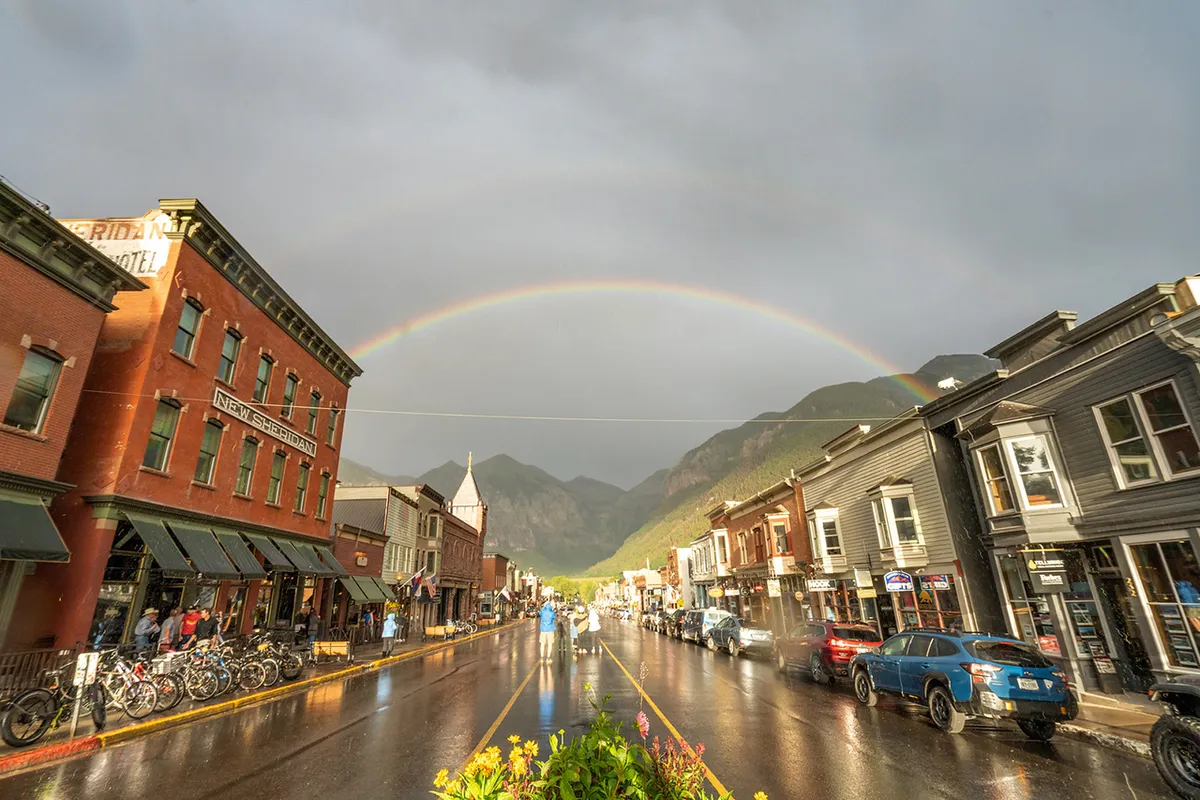 A vibrant street scene with historic buildings, a rainbow in the sky after rain, bicyclists, and bustling sidewalks. The wet street reflects the colorful buildings and the rainbow, and the presence of cafes and boutiques highlights local attractions.